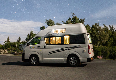 Talvor Endeavour HiTop Campervan parked in front of a forest, lights illuminating through the curtains and windows.