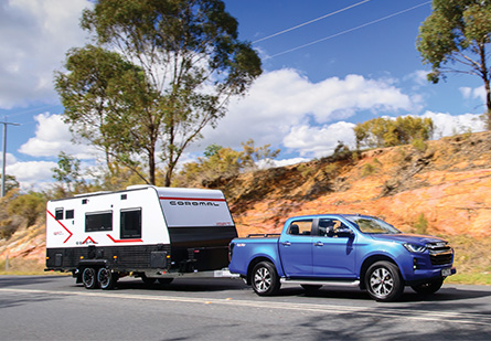 A blue ute tows a Coromal Thrill Seeker caravan on a rural Australian road. The caravan is ready for adventure, with a small hill, a tree, and blue skies with clouds in the background.