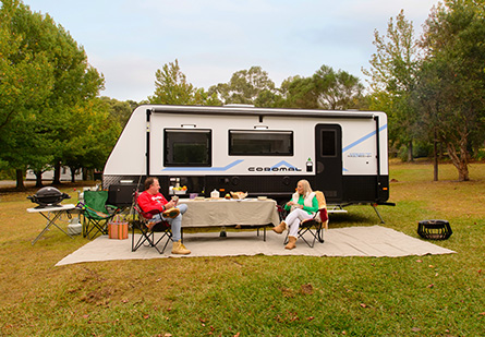 An older couple enjoys a relaxing camping setup in front of their Coromal Soul Seeker caravan. The lush green campground is surrounded by trees, creating a serene and peaceful atmosphere.