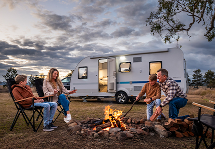 A family of four enjoying a campfire in front of their Adria Altea caravan. The scene features a cloudy sunset and camping chairs, creating a cozy and relaxing atmosphere.