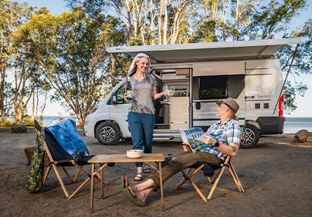 Older couple relaxing in front of an Adria Twin Campervan with an outdoor camping table and chair setup.