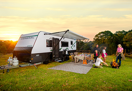 A family gathers around a campfire in front of their Coromal Adventure Seeker caravan. They enjoy the warmth of the fire and the beauty of a vibrant sunset in the background.