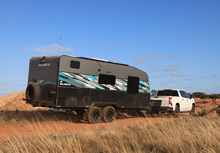 A white ute tows a Majestic Caravans Navigator caravan along a dusty rural road. Tall, dry grass lines the path, creating a sense of wild beauty and adventure.