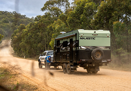 A white and blue ute tows a Majestic Caravans Trail Blazer caravan on a dusty dirt road. Trees line the path, creating a sense of secluded adventure.