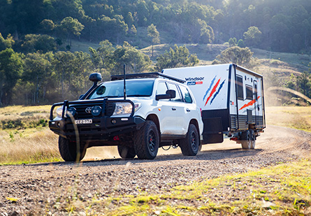 Windsor Wild Caravan being towed behind a white 4WD SUV on a winding dirt road. Hills and trees in the background.