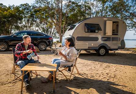 A couple enjoying a hot beverage at a campsite, with their Adria Action caravan being towed by a black SUV. The scene features a waterfront backdrop.