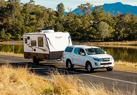 Windsor Genesis Caravan being towed behind a white ute on a quiet road by a large body of water. Trees in the background, tall grass in the forefront.