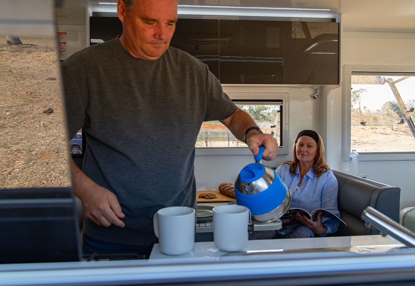 Man and woman enjoying a cup of tea inside a caravan, seated comfortably and engaging in conversation.