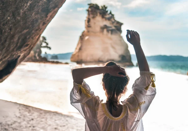 Woman with back to the camera looking at the beach and a tall rock 