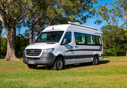 Winnebago Bondi Campervan parked on green grass in front of a trees, clear blue skies in background.