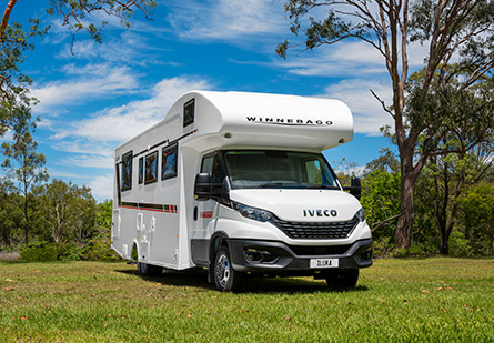 Winnebago Iluka Motorhome parked on green grass in front of a trees, clear cloudy blue skies in background.