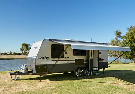 A Design RV caravan is parked on a grassy area overlooking a body of water. The clear blue sky and calm water create a serene setting for a relaxing vacation.