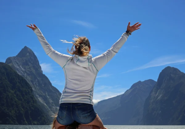 Woman on the shoulders of a person with her arms out looking at the lake and mountains of Milford Sound.