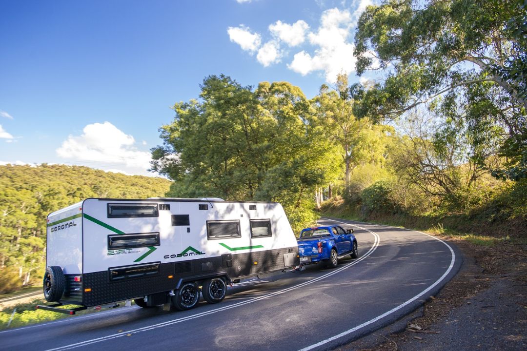 Coromal Adventure Seeker caravan being towed by a blue ute on a scenic road, surrounded by trees and hills under a clear blue sky.