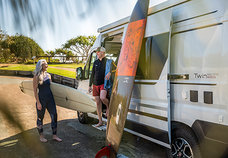 A happy senior couple stand beside their campervan in a beachfront carpark, surfboard leaning against it, ready for a day of surfing.