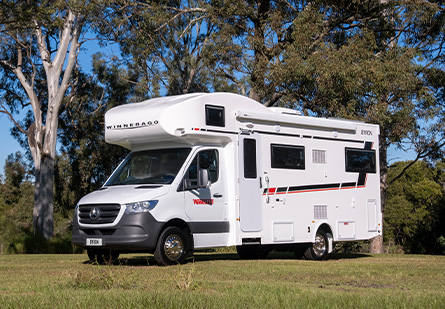 Winnebago Byron Motorhome parked on green grass in front of a trees, clear blue skies in background.