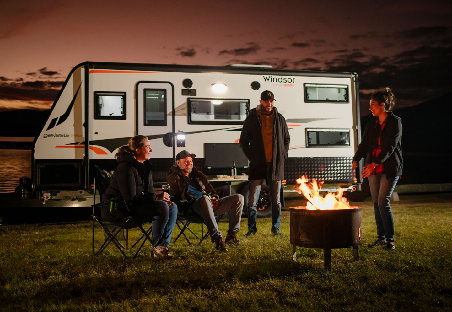 Family gathered outside around a bushfire, with their caravan visible nearby, enjoying a warm and cozy outdoor experience.