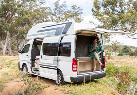 A KEA Navigator campervan is parked in a secluded bushland campsite. A woman sits in the back, enjoying a beverage, surrounded by the tranquility of nature.