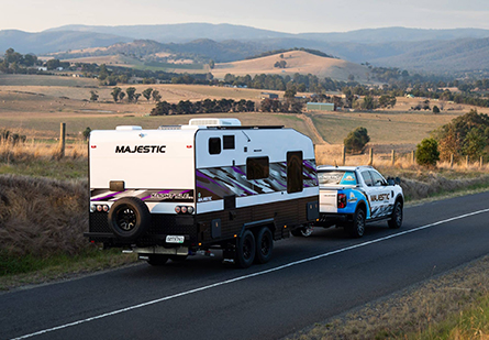 A white and blue ute pulls a Majestic Caravans Knight SLE caravan along a picturesque country road. The caravan, gleaming in the sun, promises a comfortable and luxurious adventure.