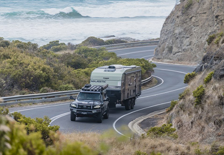 A black 4WD ute pulls a Majestic Caravans caravan along a winding road. Mountains loom on the right, while trees line the left, creating a scenic backdrop for the journey.