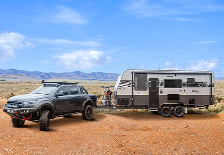 A Design RV caravan is towed by a black ute on a red dirt road, ready for off-road adventures. The rugged terrain, mountainous backdrop, and clear blue skies create a sense of freedom and exploration.