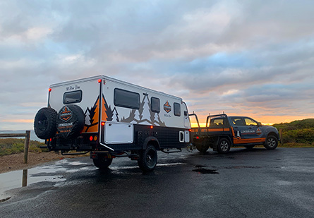 A Lumberjack MtBawBaw behing towed behind a black and orange SUV, parked on a beach carpark in front of a picturesque sunset.