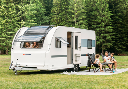 A family of five camps with their Adria Adora caravan. Dad and two kids relax outside, while mom and another child peek from the window. Lush trees and a green campsite surround them.