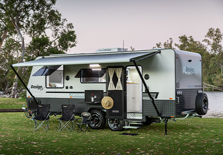 A Design RV caravan is set up on grassy ground near a body of water. Camping chairs are positioned under the awning, and a hat hangs on the front door, suggesting a relaxing and enjoyable stay.